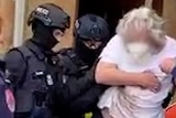 A man is held by two police officers in bullet-proof vests and balaclavas.