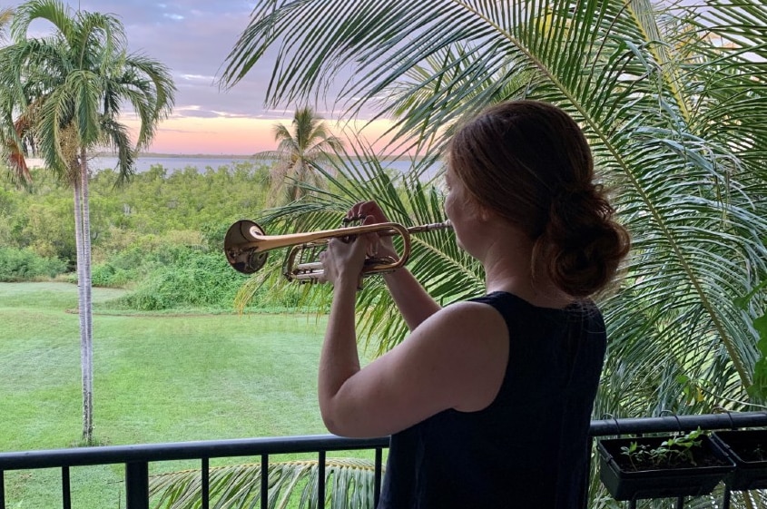 Dr Sarah Lynar plays a trumpet on her Darwin balcony overlooking a tropical garden and distant water.