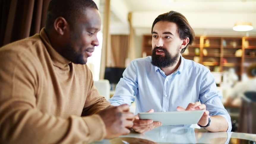 two men sitting at a table, one man talking to the other who is looking down