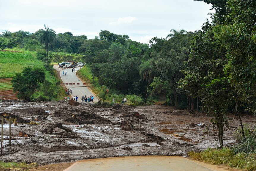 A large wide mud slide looking material covers a road at the bottom of a small valley or dip, small group of people watches on