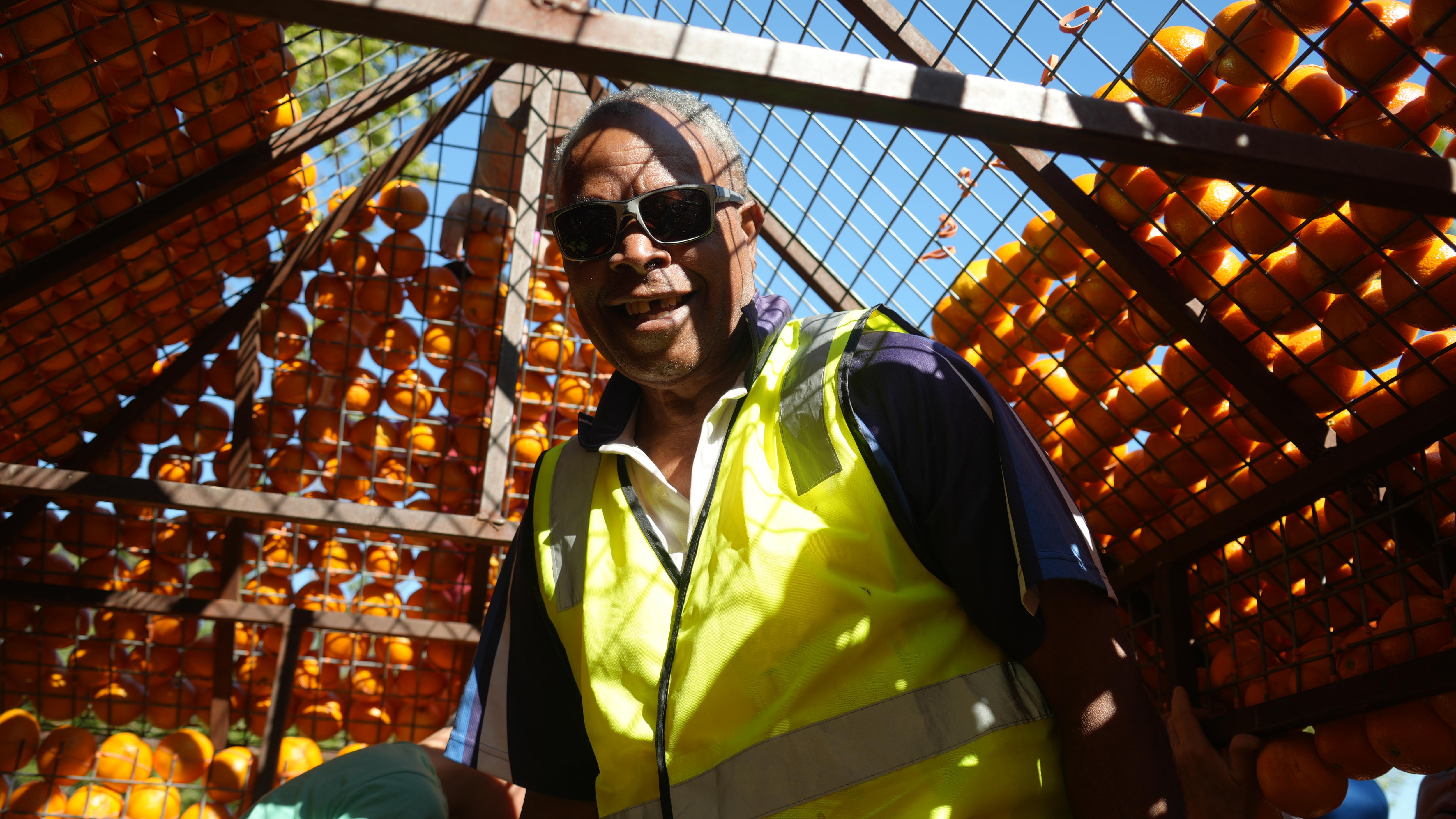 A smiling man in sunglasses and a high-vis vest stands in a replica of a house adorned with oranges.