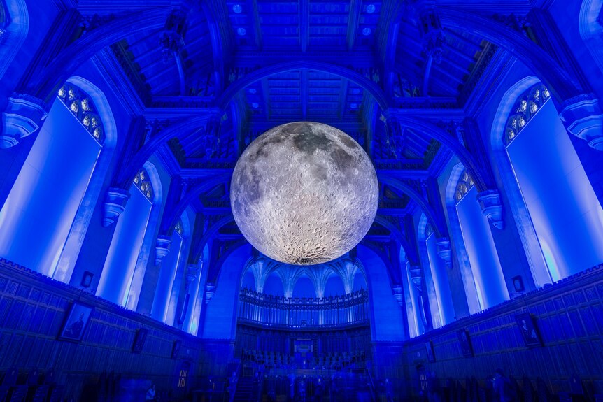 A large replica of the moon appears to be floating in a large hall bathed in blue light.