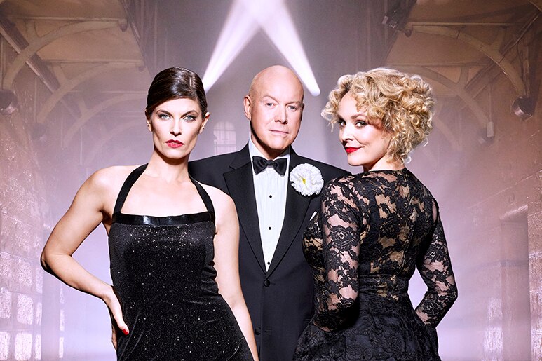 A brunette white woman and a blonde white woman in tight black dresses stand either side of a white bald man in a tuxedo