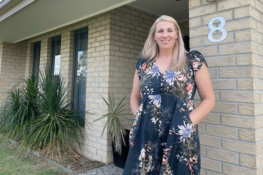 A woman in a blue floral dress stands outside her house