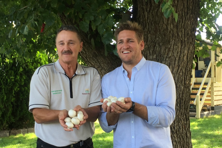 Both men smiling as they stand under a tree holding white button mushrooms in their hands.