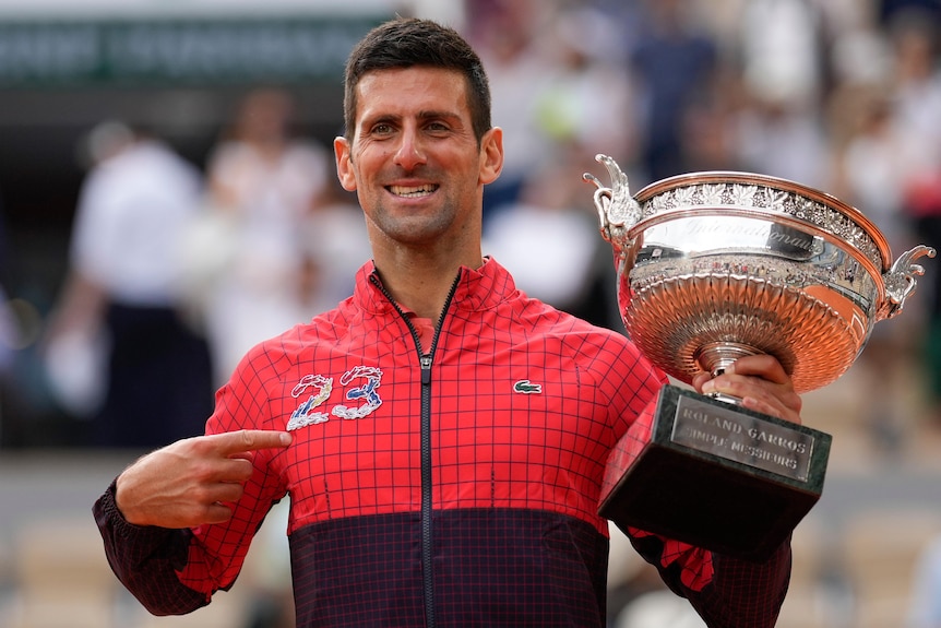 Tennis star Novak Djokovic smiles as he holds the French Open trophy while wearing a jacket with "23" on it.