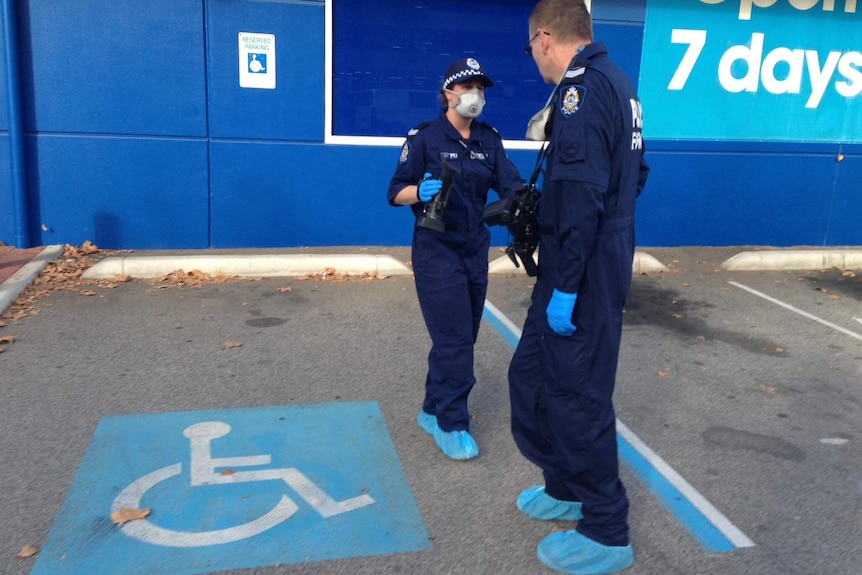 Two police forensic officers in blue jumpsuits and facemasks talk outside a stationery shop.