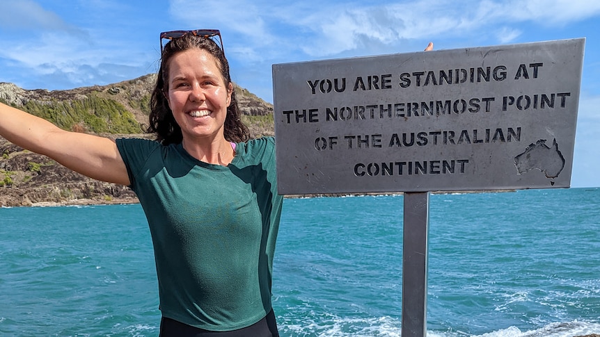 A smiling woman stands by a sign with the sea and an outcrop of  land behind her