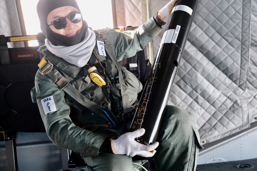 Researcher Pat Wongpan in thermals holding a black cylinder in the back of a helicopter.