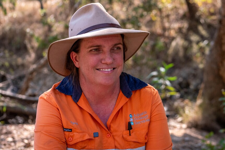 A woman in a high vis orange shirt and a hat sits smiling on a riverbank