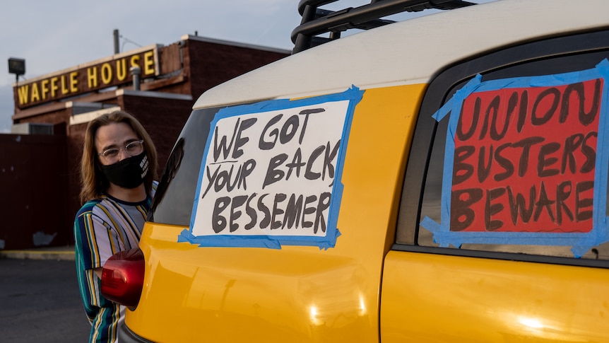 A man with glasses peeks out behind a yellow car bearing homemade banners reading, "We've got your back, Bessemer"