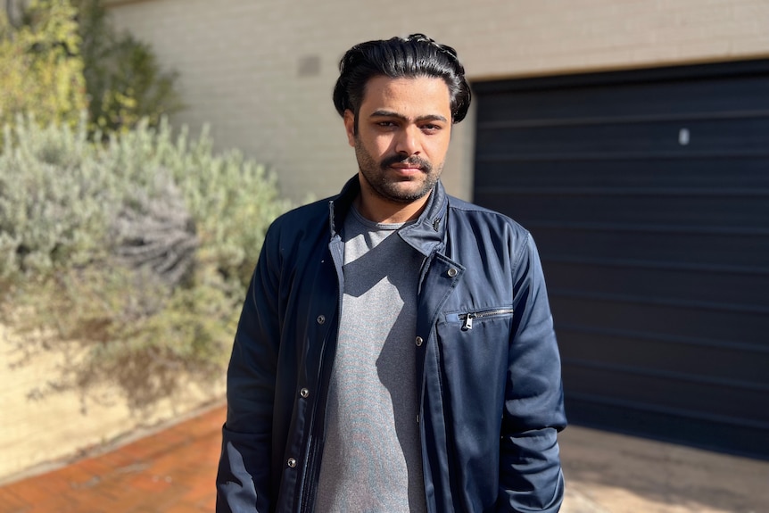 A man in a leather jacket stands outside a house looking serious.