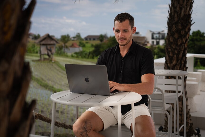 A young man in shorts looks at his laptop at an outdoor cafe