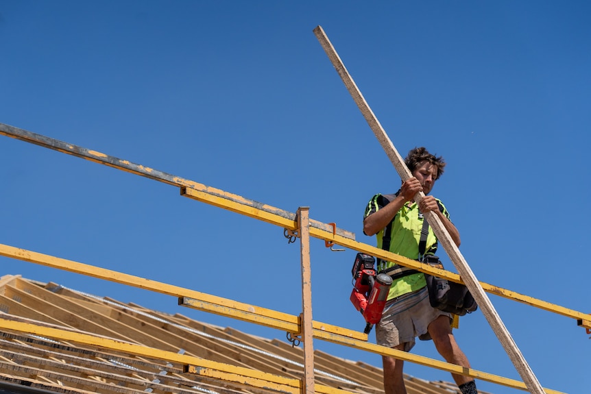 a man on a roof lifts a wooden plank over scaffolding. 