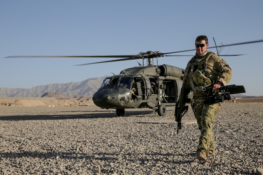 A man wearing camouflaged gear walks in front of a helicopter.
