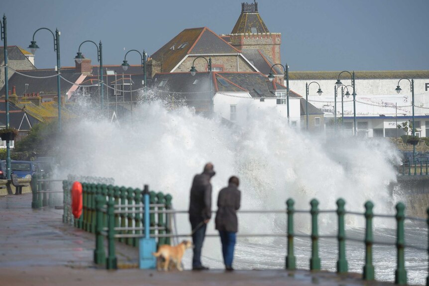 A couple watch waves break on the sea wall at Penzanze, England. A huge wave obscures the buildings built near the sea wall.