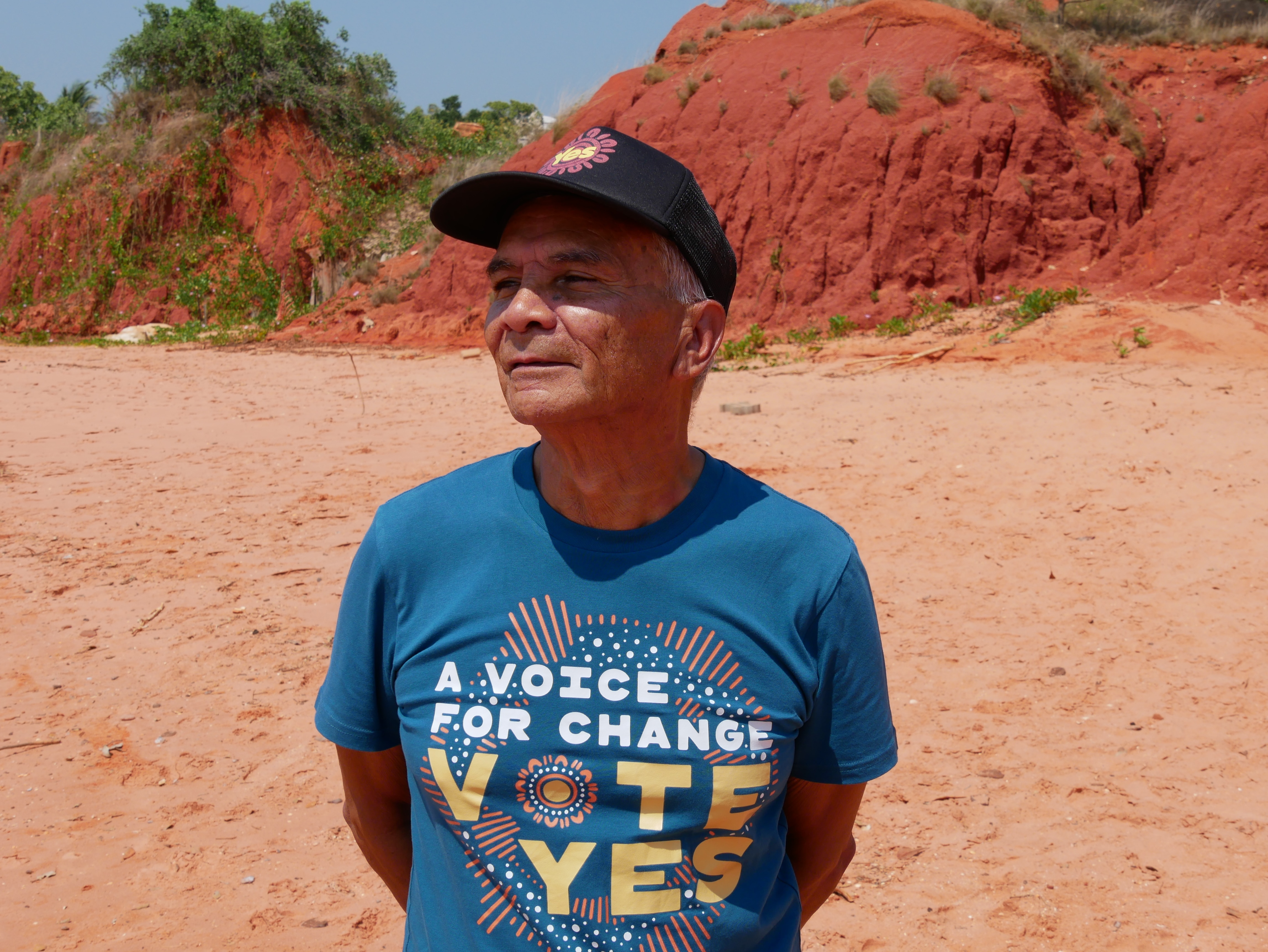 A man standing on a beach wearing a shirt that reads "a voice for change, vote yes".