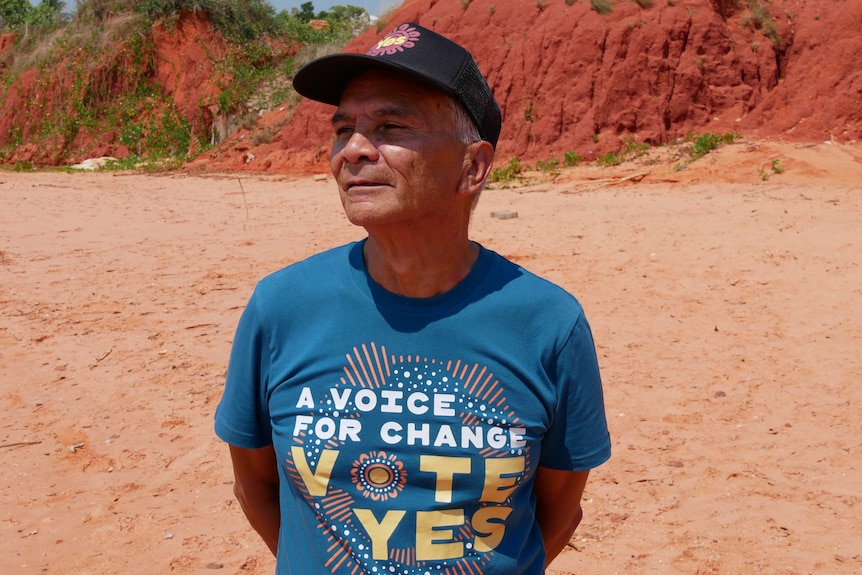 A man standing on a beach wearing a shirt that reads "a voice for change, vote yes".