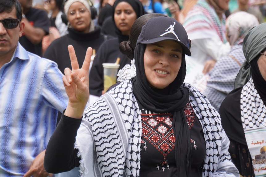 a woman at a pro-palestinian rally in perth does the peace sign with her two fingers as she smiles at the camera