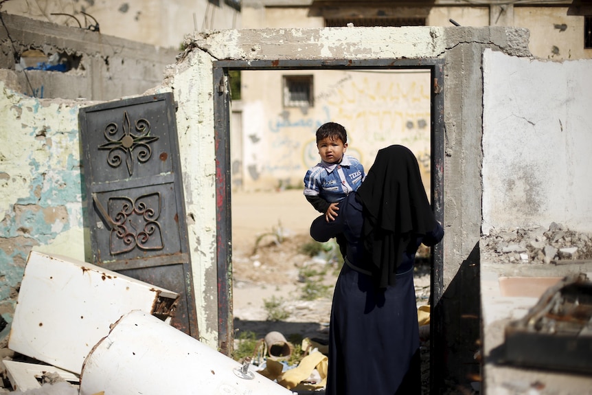 A woman wearing dark clothing and head covering carries a little boy through the doorway of a home in ruins