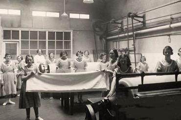 Black and white grainy image of women standing at large laundry troughs and holding sheets in a room.