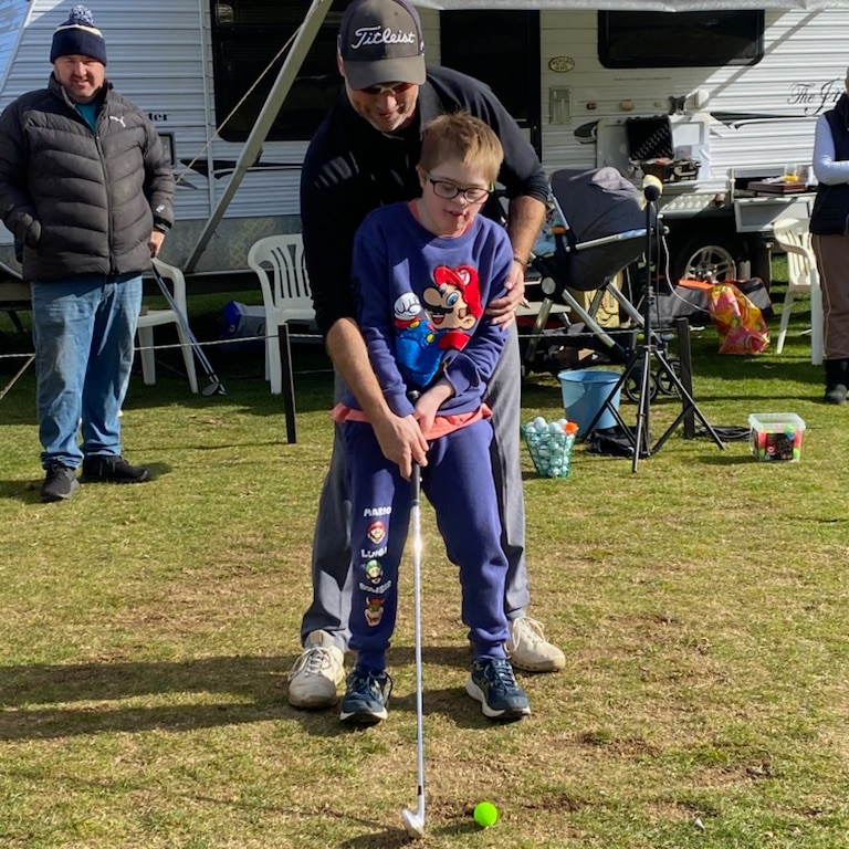 A man in a baseball cap shows a child with Down Syndrome how to swing a golf club