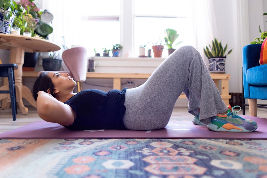 A woman lays on a mat in a living room doing a sit up while laying on a yoga mat. She wears sweat pants and shirt, and sneakers.