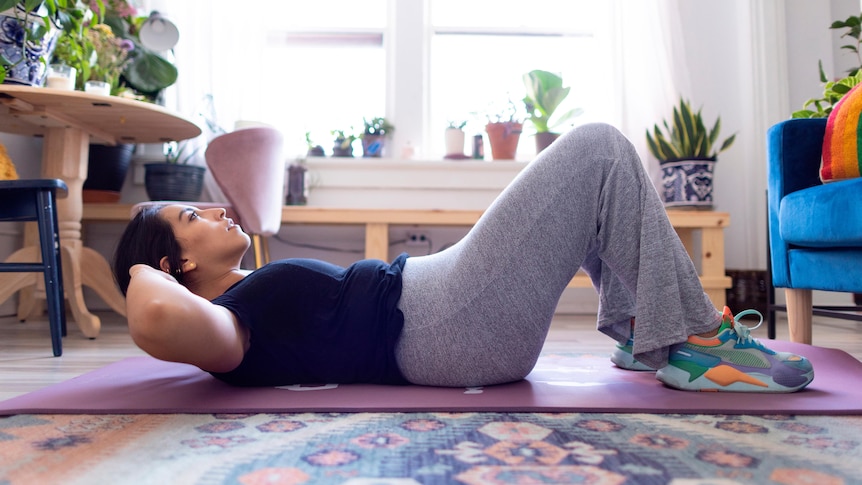 A woman lays on a mat in a living room doing a sit up while laying on a yoga mat. She wears sweat pants and shirt, and sneakers.