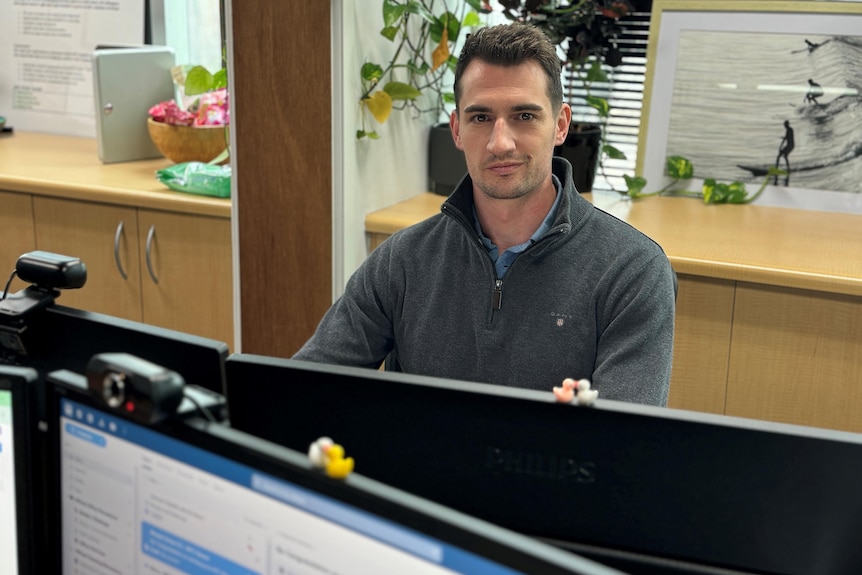A man wearing a grey half-zip pullover jumper sits at a desk behind two computer monitor screens in an office.