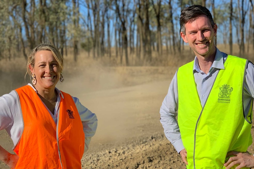 A woman and man in high visibility vests stand on the side of a dirt road with dust in the air behind them.