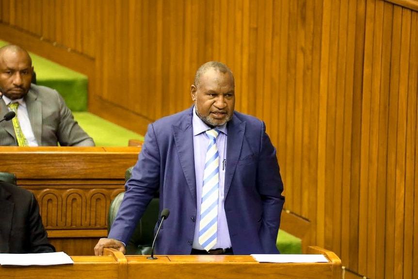 James Marape in blue suit stands in a parliamentary chamber with wooden benches and green carpet