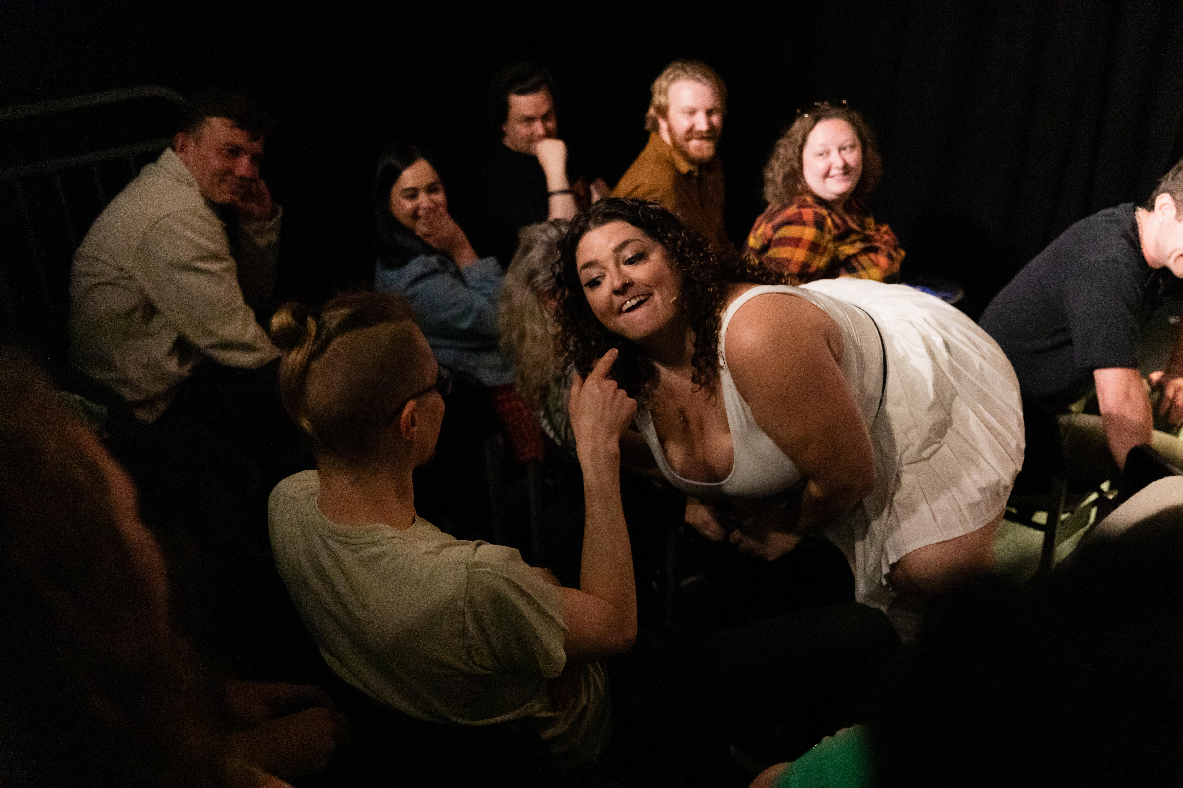 Uma mulher de vestido branco se ajoelha diante de um homem que aponta para ela, com o público sorrindo ao fundo.