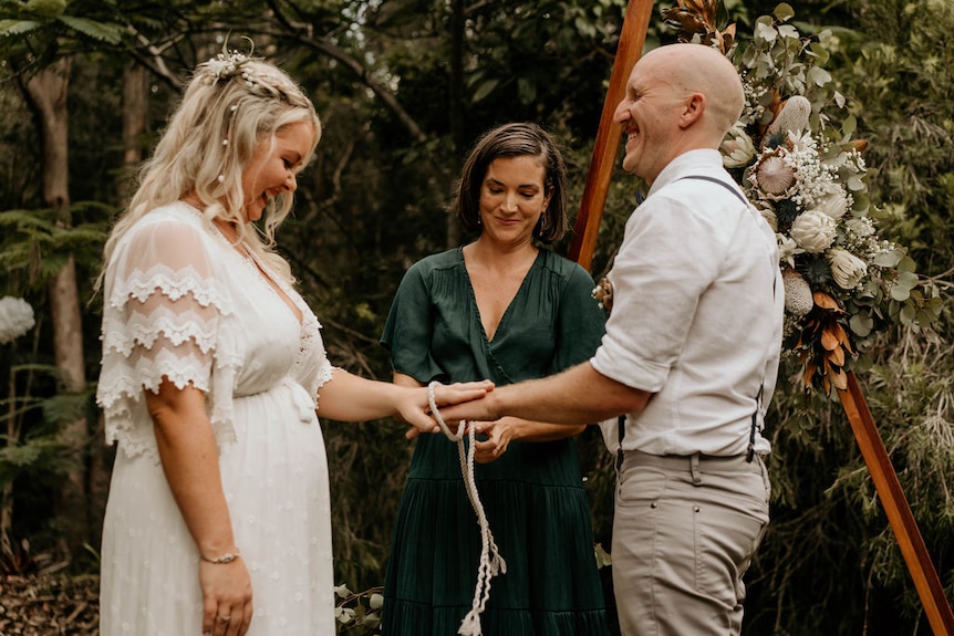 A bride and groom holding hands and celebrant between them holding a rope around their hands.
