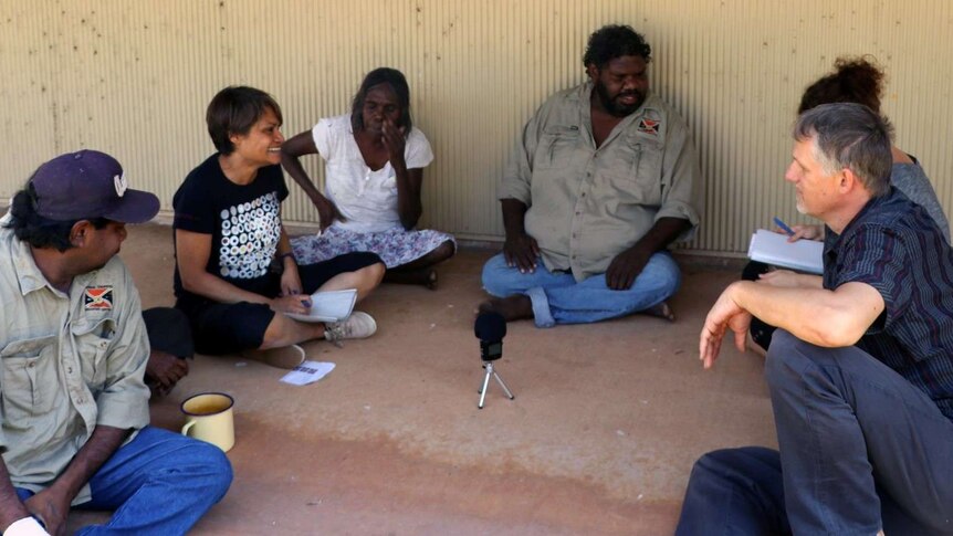 A group sit around a recording device, outside on a veranda