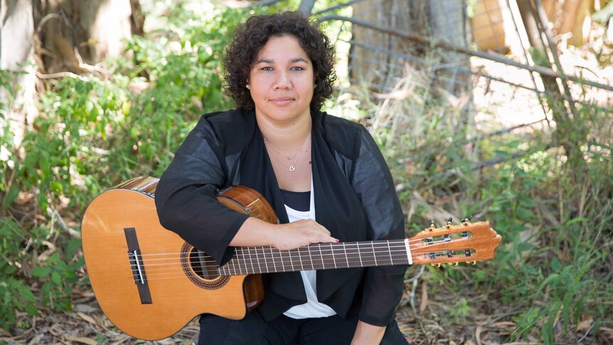 Aboriginal and Torres Strait Islander musician Jessie Lloyd holds guitar and sits on stool in overgrown garden.