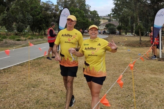 A tall man in black shorts and a yellow tee and cap branded with 'Achilles Melbourne' next to a younger woman in the same. 