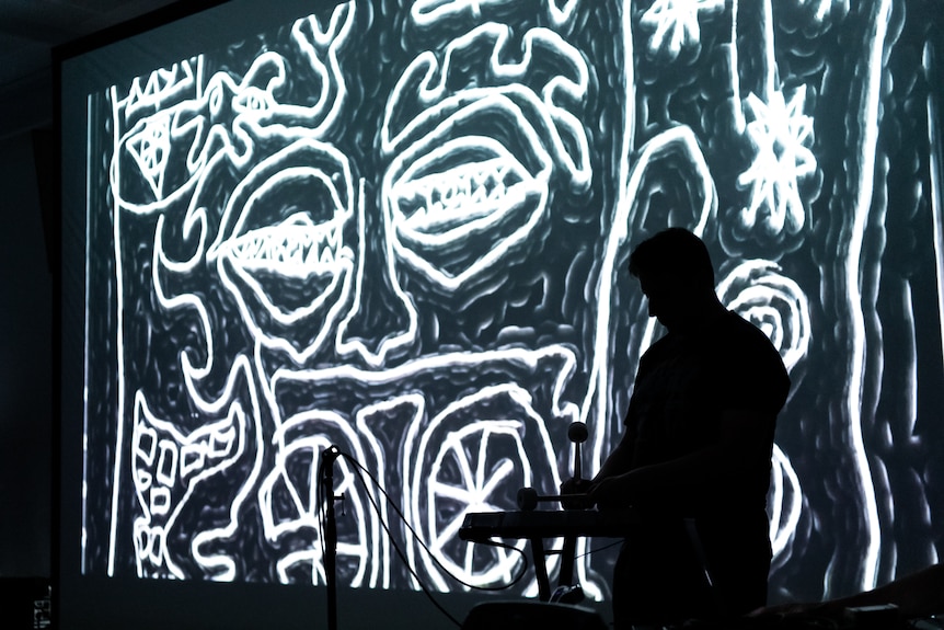 A man plays a zinc ingot silhouetted against a black and white screen