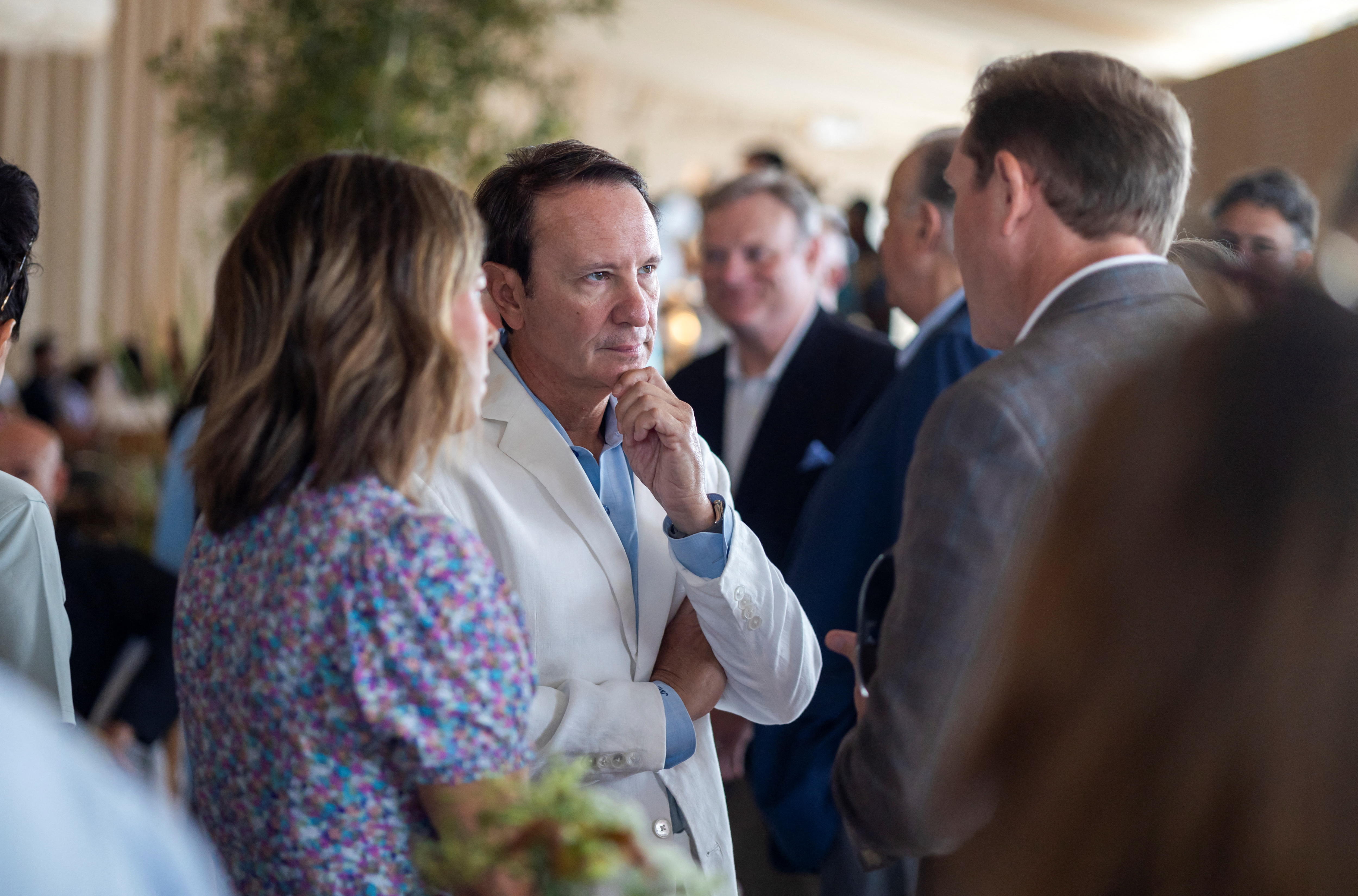 Un hombre con una chaqueta blanca y una camisa azul está hablando con otro hombre, una mujer está parada junto a él.