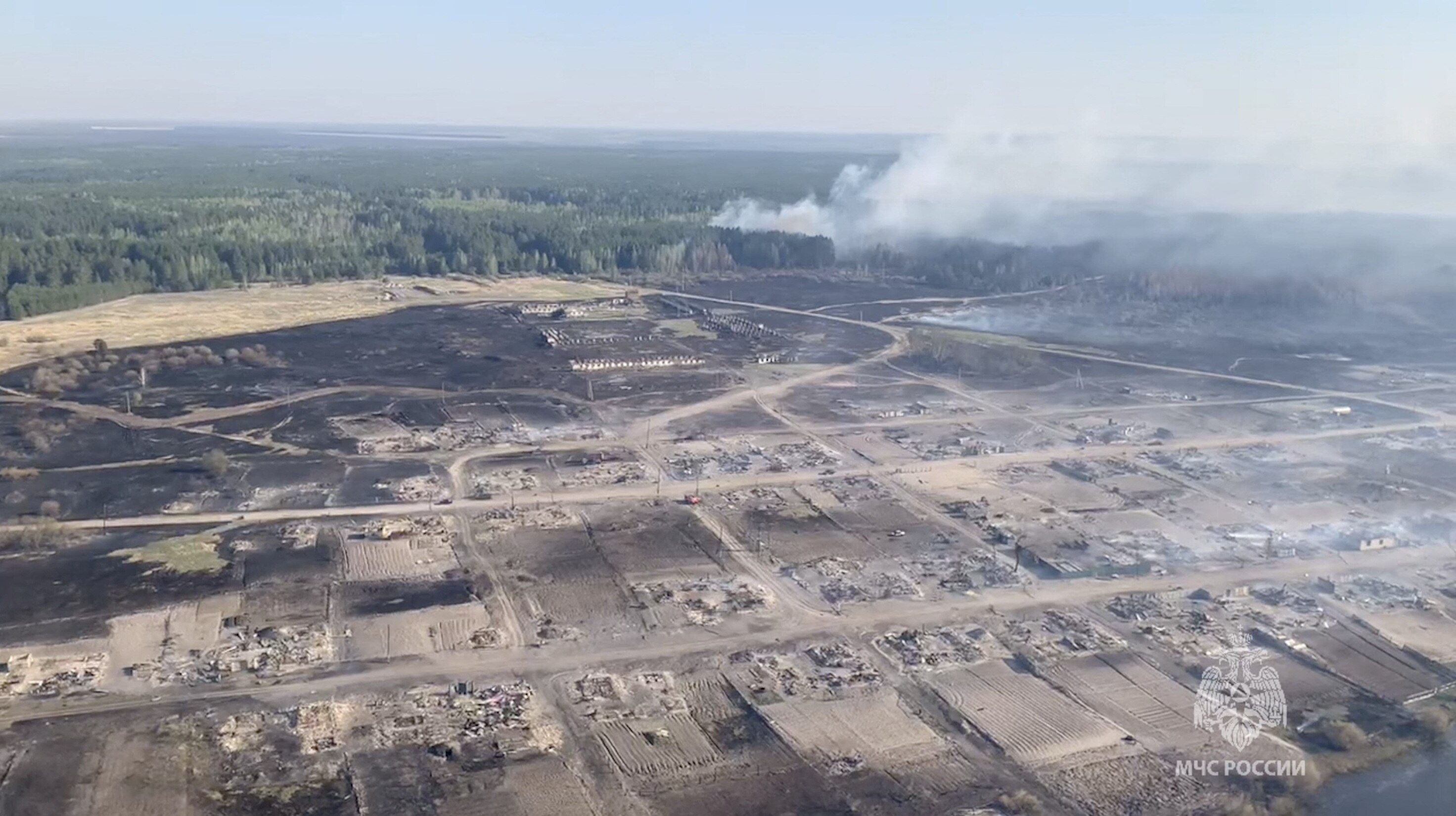 A view from a helicopter shows charred grounds and a large plume of smoke coming from a forested area.