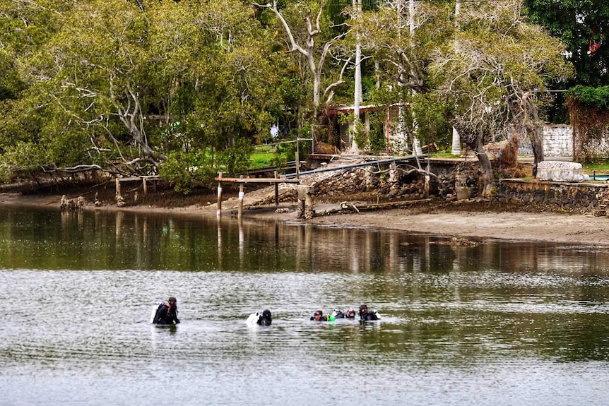 Diver waist-deep in a creek about 10 metres from the shoreline, near trees, a waterfront house and jetties