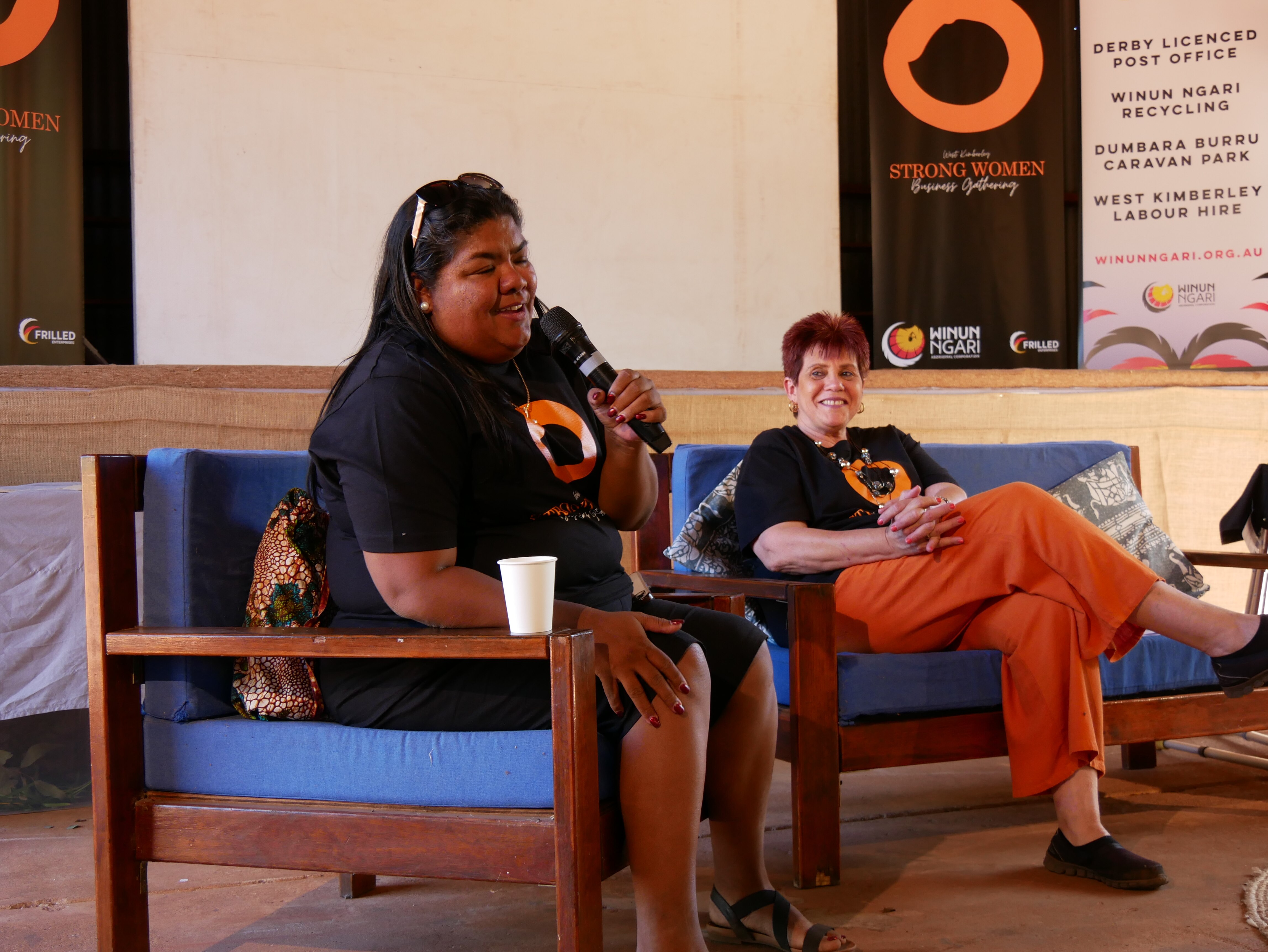 An Indigenous woman in a black shirt with long dark hair and glasses on her head holds a microphone