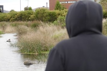 A figure clad in a dark hoodie sits in front of a lake. Identity hidden.