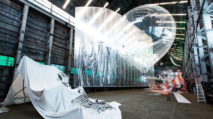 An installing of a giant clear plastic balloon, hanging reflective plastic sheeting among other things inside a warehouse space.