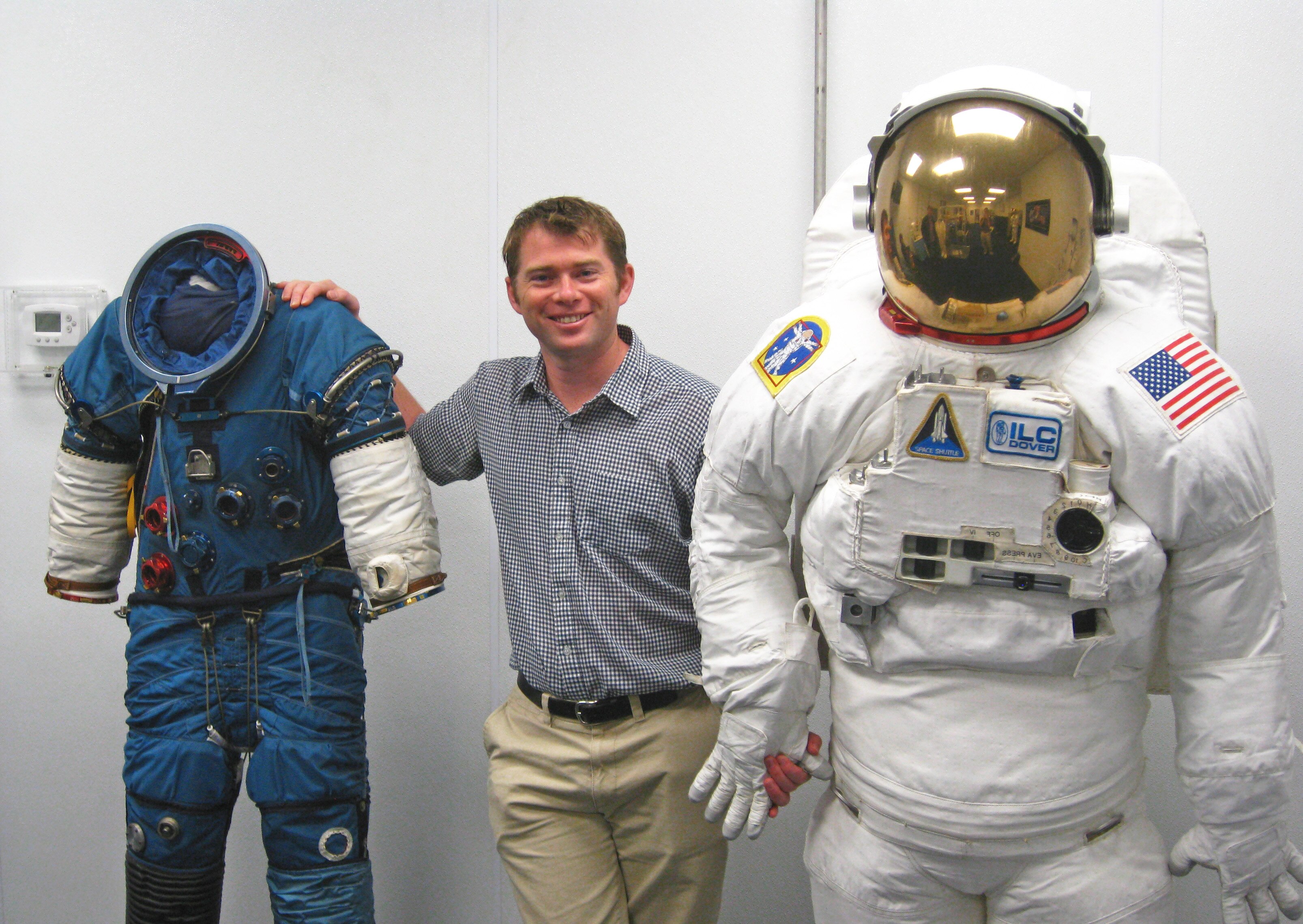 A man stands between a modern space suit and the classic Apollo-era astronaut suits smiling.