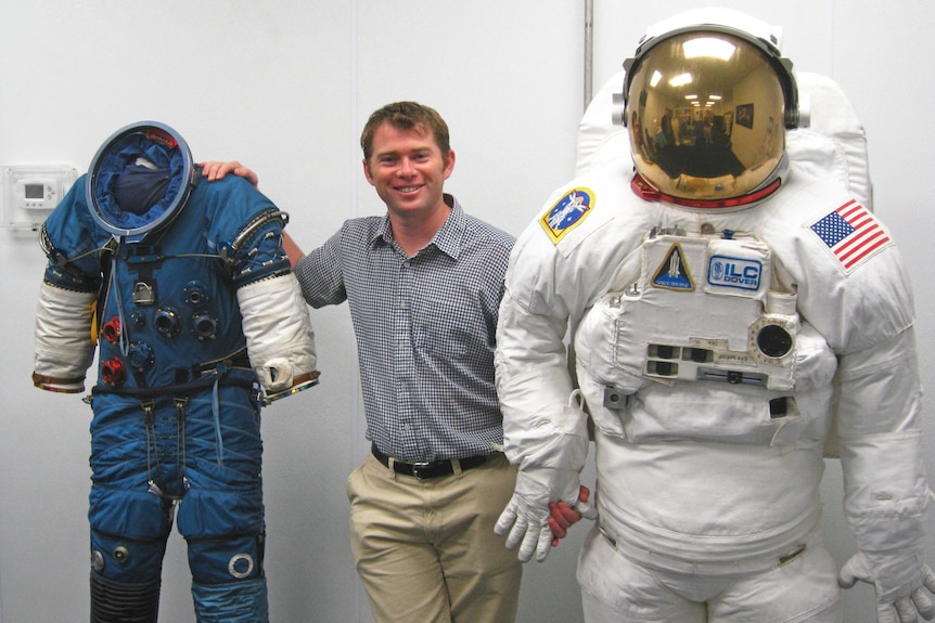 A man stands between a modern space suit and the classic Apollo-era astronaut suits smiling.