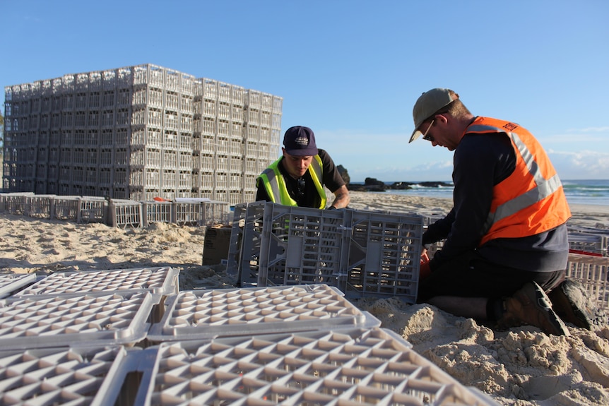 Two men on a beach constructing a sculpture made from plastic milk crates. 