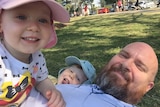 John Petrak, with his children Eleanor and JB, laying on the grass in a park on Queensland's Gold Coast.