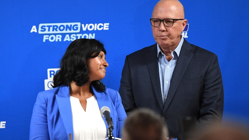 Roshena Campbell and Peter Dutton look downcast while standing on a blue stage.