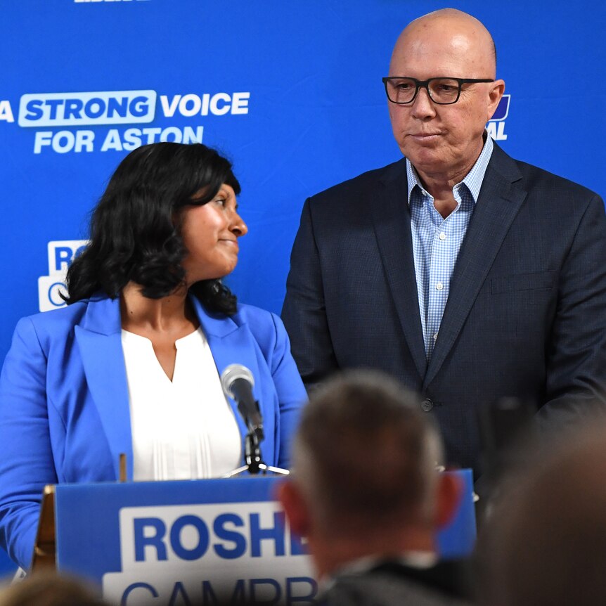 Roshena Campbell and Peter Dutton look downcast while standing on a blue stage.