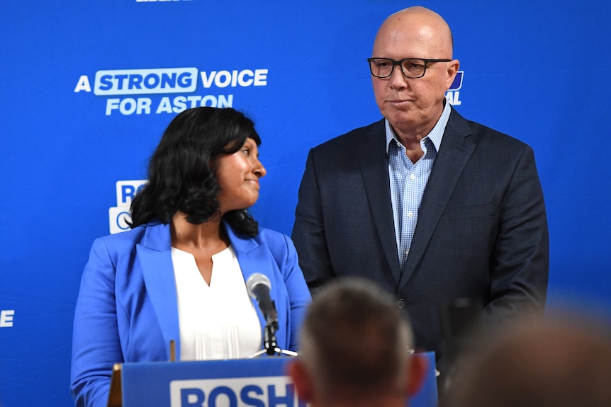 Roshena Campbell and Peter Dutton look downcast while standing on a stage with a blue Liberal poster behind them.